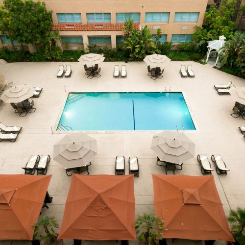 Aerial view of a hotel pool area with a rectangular blue pool, multiple lounge chairs, umbrellas, and surrounding greenery, balconies above.