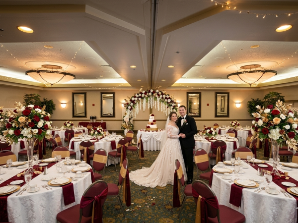 A wedding reception with a couple standing under a floral arch, round tables draped in white and burgundy, and elegant chandeliers in a banquet hall.