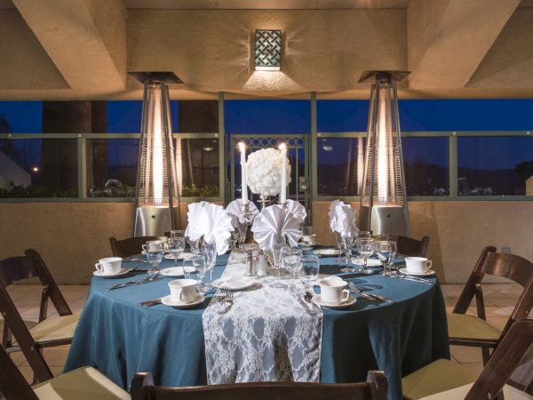 A round, teal-draped dinner table set for four with white floral centerpieces and place settings under warm patio heaters, in an evening venue.