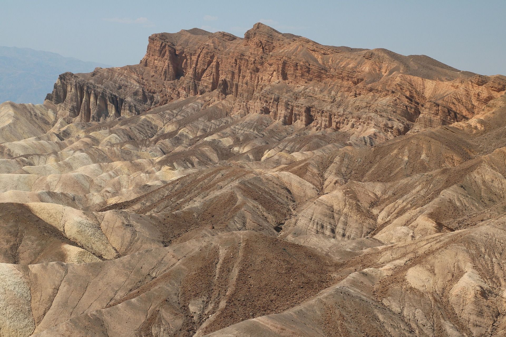 A rugged desert mountain ridge with steep, layered rock faces and deep gullies under a pale blue sky.