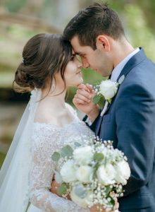 A bride and groom share a tender moment, noses almost touching, at a wedding with a bouquet and soft greenery in the background.