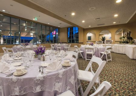 A banquet hall set for a formal event: round tables with white linens, chairs, place settings, centerpieces, and buffet area in the background.